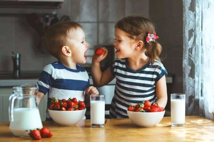 Two chidren sitting at a table. The little girl is giving the little boy a strawberry. They have bowls of strawberries and glasses of milk on the table in front of them.