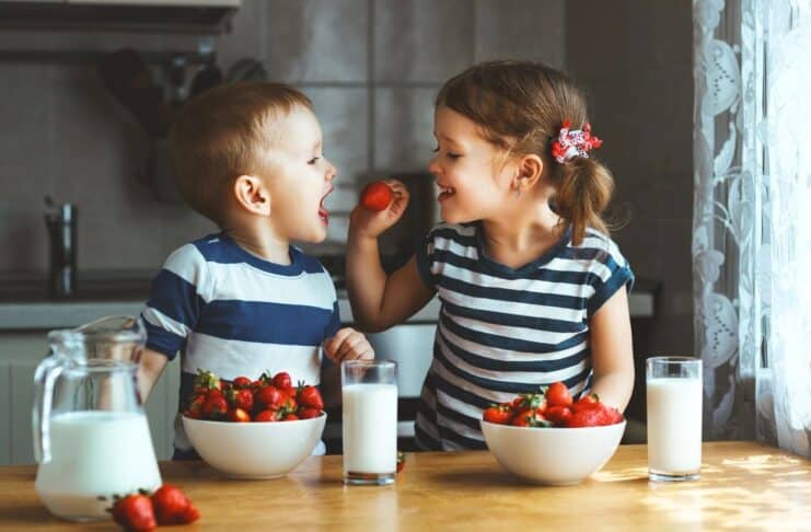 Two chidren sitting at a table. The little girl is giving the little boy a strawberry. They have bowls of strawberries and glasses of milk on the table in front of them.